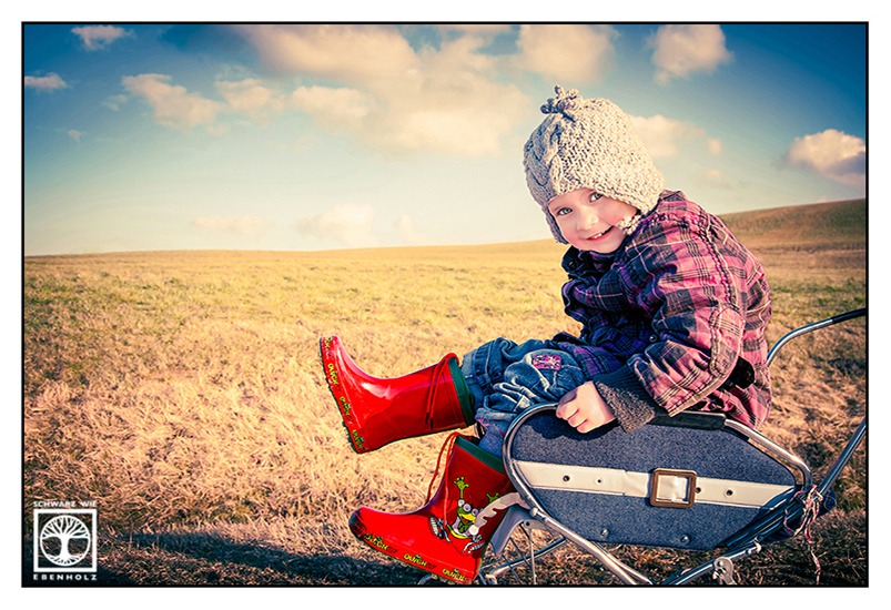 Outdoor photoshoot child in the countryside: This photo shows a little girl sitting in an old-fashioned doll's pram in front of a barren field and a blue sky with some clouds. She is wearing a grey bonnet, a pink checkered jacket, blue jeans and red rubber boots. You can see a green frog on her left boot.