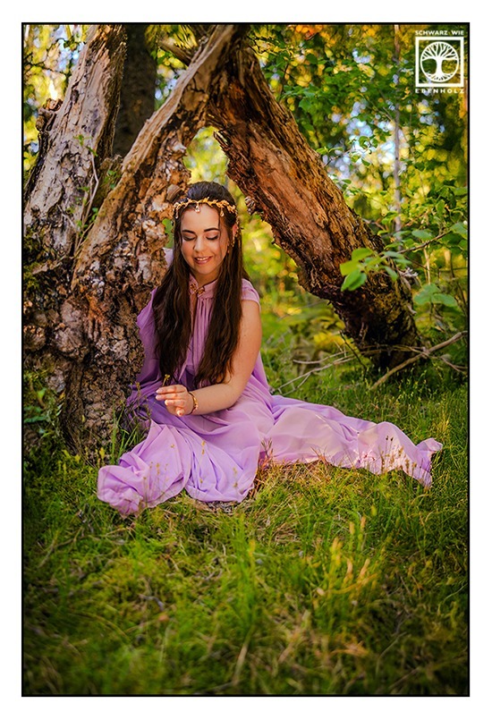 Fantasy Fotoshooting: Göttin Aphrodite sitzt im Wald unter einem abgebrochenen alten Baum. Sie trägt ein fliederfarbenes Kleid und goldenen Schmuck in Form von Blättern im Haar. Ihr Blick ist auf eine Blume gerichtet, die sie gerade pflückt.