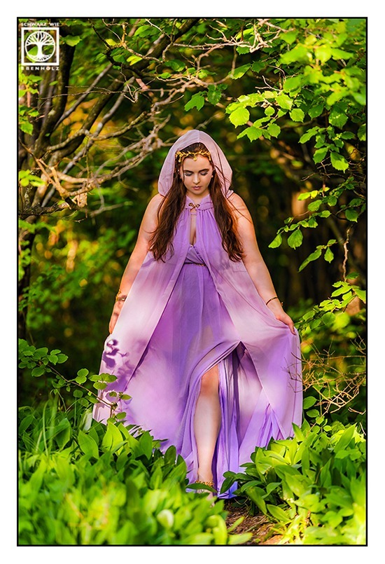 Fantasy Fotoshooting: Göttin Aphrodite läuft durch den Wald, umgeben von den grünen Blättern des Maiglöckchens. Sie trägt ein fliederfarbenes Kleid und goldenen Schmuck in Form von Blättern im Haar. Auf dem Kopf ist die Kapuze ihres farblich passenden Umhangs, den sie mit ihren Händen ein wenig hochhält. Ihr Blick ist auf den Boden gerichtet.