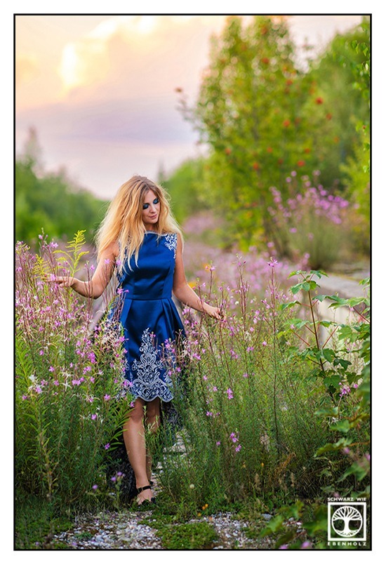 Fotoshooting Natürlich: Eine Frau mit langen blonden Haaren und elegantem knielangem blauen Kleid mit weißen Stickereien läuft durch eine Blumenwiese. Ihre Finger streichen dabei über die rosa Blumen. Ihr Blick ruht auf ihnen. Das Bild ist Teil der Serie "Blue is just a New Beginning" (deutsch: "Blau = Traurigkeit ist nur ein neuer Anfang". Das Einzelbild heißt "Gaining strength" (deutsch: "An Stärke gewinnen").