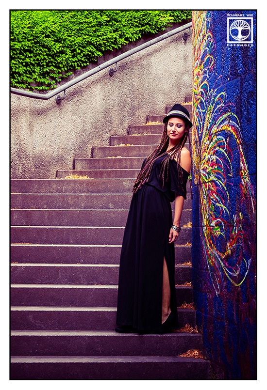 Boho Fotoshooting / Alternative Fotoshooting: Eine junge Frau steht neben einem bunten Graffiti eines Vogels auf einer Treppe. Oberhalb des Geländes wächst Efeu. Ihr Oberkörper neigt sich in Richtung der Wand, so dass es aussieht als hätte sie Flügel. Ihre Arme hängen hinab. Sie blickt nach unten. Auf ihrem Kopf ist ein schwarzer Hut mit buntem Band im Ethno-Look. Sie hat lila und dunkelbraune Braids, die ihr bis zur Hüfte reichen und trägt ein langes, schwarzes Kleid mit kurzen Ärmeln. An ihrem Armgelenk sind silberne Reifen.