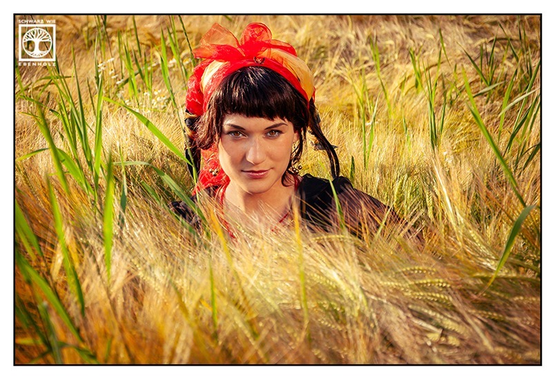 Fantasy Fotoshooting Faun / Fauna: Eine Frau mit schwarzem Haar und transparenter schwarzer Bluse sitzt in einem Kornfeld. Sie trägt einen Kopfschmuck, der wie zwei Hörner aussieht, die mit rotem und gelbem Hüll umwickelt wurden. Sie schaut ernst und blickt uns direkt in die Augen.