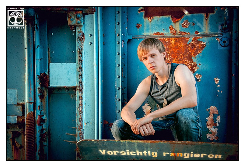 Outdoor Fotoshooting: Ein junger Mann mit blonden kurzen Haaren mit ernsten Gesichtsausdruck sitzt in der Hocke mit darauf abgestützten Armen vor einem blauen Eisenbahnwaggon mit orange-roten Rostflecken. Er trägt ein graublaues Achselshirt und eine blaue Jeans.