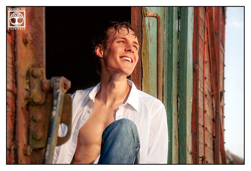 Outdoor Fotoshooting Mann: Ein junger Mann sitzt in einem rostigen Eisenbahnwaggon. Er trägt ein offenes weißes Hemd und eine blaue Jeans. Sein kurzes Haar ist nass und verstrubbelt. Er schaut nach oben in die von dir aus rechte Ecke und lacht.