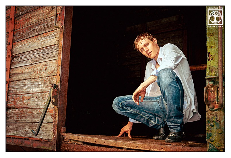 Outdoor Fotoshooting: Ein junger Mann mit nassen dunkelblonden Haaren sitzt in der Hocke im Inneren eines geöffneten Eisenbahnwaggons. Das Bild ist leicht von der Seite aufgenommen. Er trägt ein weißes Hemd, eine blaue Jeans und schwarze Schuhe.