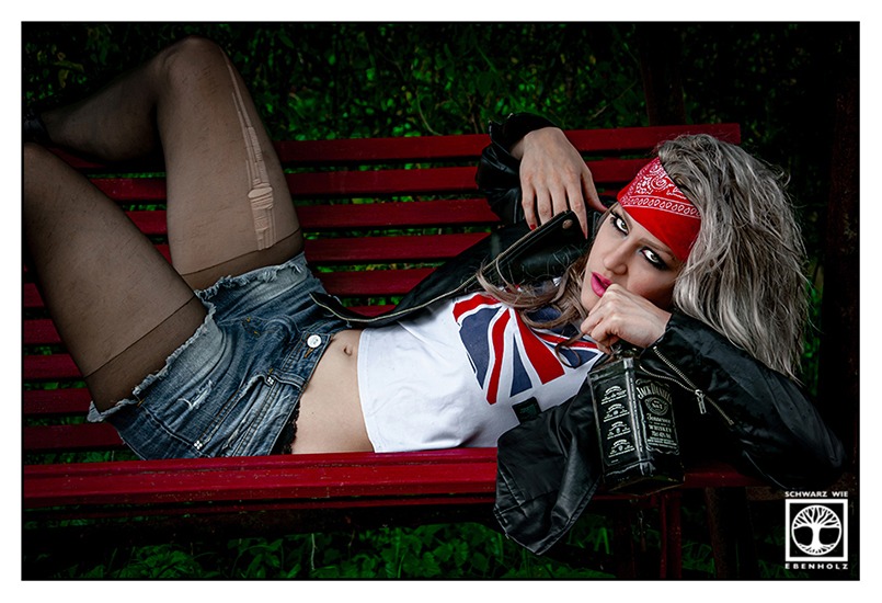 Outdoor Fotoshooting im Grunge Look: Eine Rockerbraut mit grauen langen Haaren, bauchfreiem weißen Shirt mit britischer Flagge, Lederjacke, rotem Stirnband, kurzen Bluejeans und zerrissener Strumpfhose liegt provokant auf einer roten Bank. Sie blickt auffordernd in die Kamera und versucht gerade, eine Whiskeyflasche mit dem Mund zu öffnen.