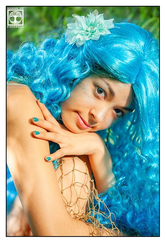 Fantasy Fotoshooting: Das hier ist ein Beauty Portrait einer schlanken jungen Frau in türkisem Bikini und mit langen lockigen blauen Haaren und weißer Stoffblume im Haar. Sie sitzt in der Hocke und hat ihre Beine an den Körper gezogen. Ein Fischernetz bedeckt dürftig ihre Beine. Nur Kopf, Arme und Knie sind zu sehen. Sie blickt uns an.