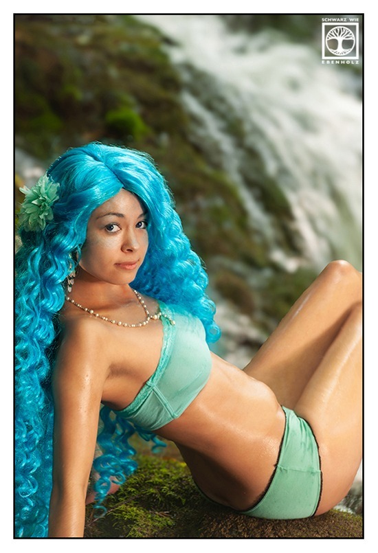 Fantasy Fotoshooting: Eine schlanke junge Frau in türkisem Bikini und mit langen lockigen blauen Haaren und weißer Stoffblume im Haar sitzt auf einem moosigen Felsen vor einem Wasserfall. Sie blickt uns über die Schulter direkt an. Arm und Beine sind angeschnitten.