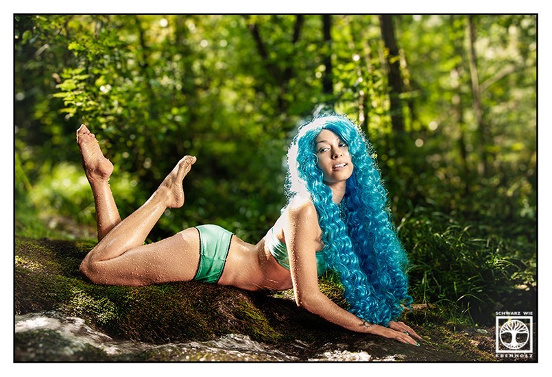 Fantasy Fotoshooting: Eine schlanke junge Frau in türkisem Bikini und mit langen lockigen blauen Haaren liegt mit dem Bauch auf einem moosigen Felsen neben einem Wasserfall im Wald. Ihre Beine sind auf verschiedenen Höhen angewinkelt. Sie stützt sich auf ihre Unterarme und lächelt selbstbewusst. Ihr Blick ist nach hinten gewandt.