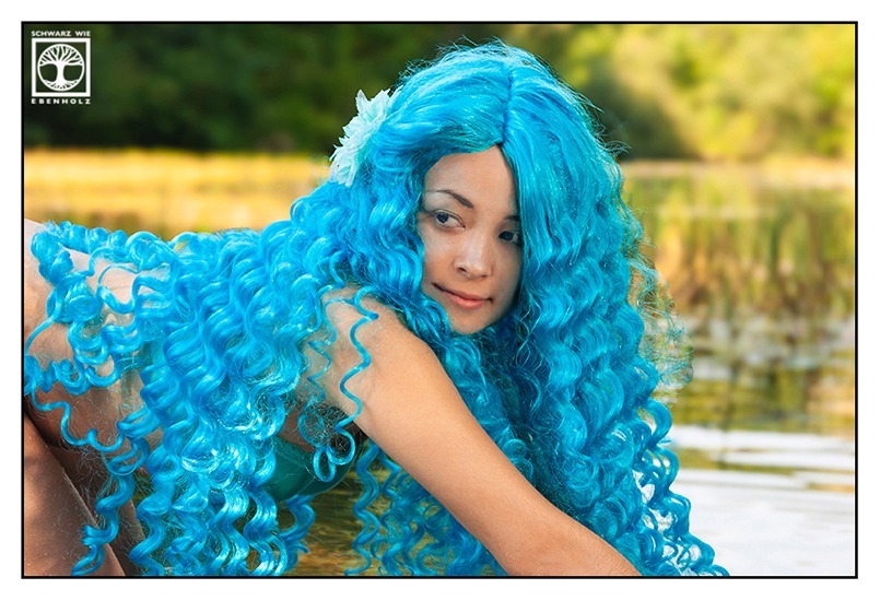 Fantasy Fotoshooting Wassernymphe: Eine schlanke junge Frau in türkisem Bikini und mit langen lockigen blauen Haaren und hellblauer Stoffblume im Haar befindet sich auf allen Vieren und blickt über ihre rechte Schulter zurück. Der Betrachter sieht ihre rechte Seite, aber nur einen Anschnitt der Arme und Beine. Im Hintergrund ist ein natürlicher See.