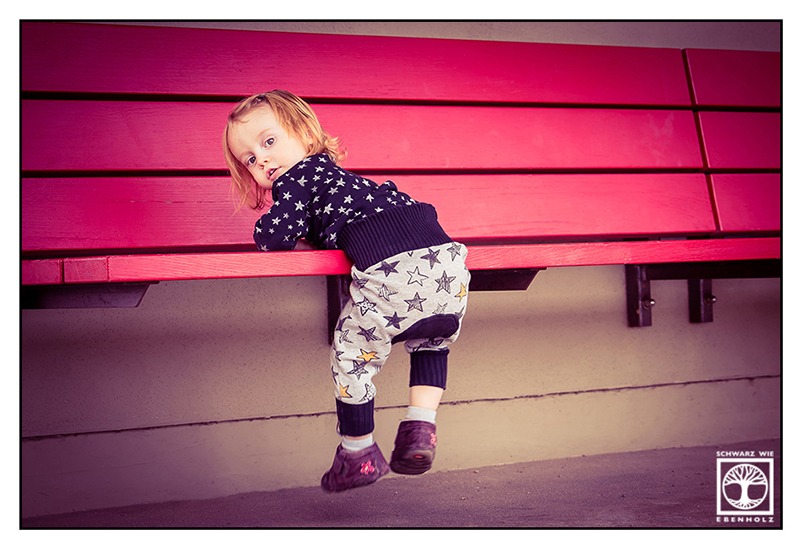 Outdoor Fotoshooting im Park: Das Foto zeigt ein kleines Kind mit halblangen blonden Haaren, das halb auf der Sitzfläche einer hohen roten Bank hängt. Die Füße baumeln in der Luft. Sie stecken in lila Schuhen mit je einer rosa Blume darauf. Das Kind trägt eine graue Hose mit schwarzen und gelben Sternen und einen schwarzen Pulli mit weißen Sternen. Es blickt über die uns zugewandte Schulter zu uns.