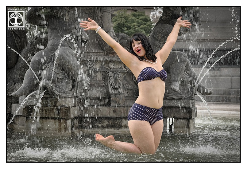 Outdoor Fotoshooting im Vintage Look: Im marineblauen Polkadot Bikini springt eine Frau mit schwarzen halblangen Locken durch einen Brunnen mitten in der Innenstadt von Stuttgart und hat sichtlich Spaß daran. Um sie herum spritzt das Wasser aus den Steinfiguren.