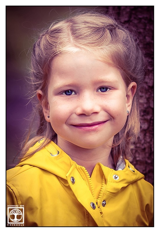 Outdoor Fotoshooting Kind im Wald: Das Halbporträt zeigt ein lächelndes blondes Mädchen mit blauen Augen im Vorschulalter. Ihre Haare sind nach hinten weg geflochten. Sie trägt eine gelbe Regenjacke und Ohrstecker. Der Titel lautet auf deutsch: "Sonne an einem regnerischen Tag."