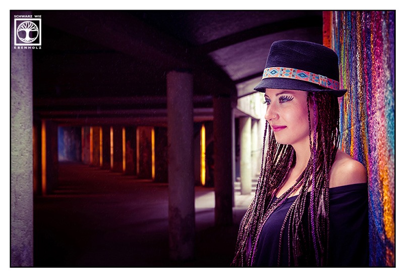 Boho Fotoshooting: Eine junge Frau steht seitlich in einer Unterführung direkt neben einem farbenfrohen Graffiti und verdeckt fast vollständig eine lichtüberflutete Treppe. Sie blickt nachdenklich in die Ferne Auf ihrem Kopf ist ein schwarzer Hut mit buntem Band im Ethno-Look. Sie hat lila und dunkelbraune Braids, die ihr bis zur Hüfte reichen und trägt ein langes, schwarzes Kleid mit kurzen Ärmeln und U-Boot-Ausschnitt.