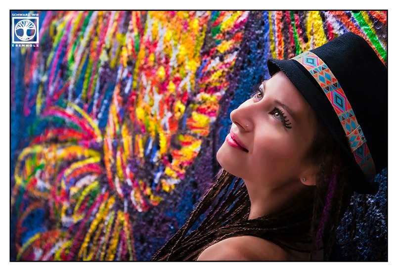 Boho Fotoshooting / Alternative Fotoshooting: Eine junge Frau steht neben einem bunten Graffiti eines Vogels und blickt lächelnd in den Himmel. Auf ihrem Kopf ist ein schwarzer Hut mit buntem Band im Ethno-Look. Sie hat lila und dunkelbraune Braids, die ihr bis zur Hüfte reichen. Das Foto zeigt nur ihren Kopf und die Schulter.
