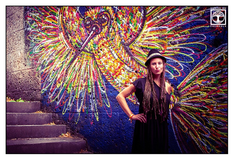 Boho Fotoshooting / Alternative Fotoshooting: Eine junge Frau steht vor einem bunten Graffiti eines Vogels, stemmt ihren rechten Arm in die Hüfte und macht einen Kussmund. Auf ihrem Kopf ist ein schwarzer Hut mit buntem Band im Ethno-Look. Sie hat lila und dunkelbraune Braids, die ihr bis zur Hüfte reichen und trägt ein langes, schwarzes Kleid mit kurzen Ärmeln. An ihrem Armgelenk sind Festivalbänder.