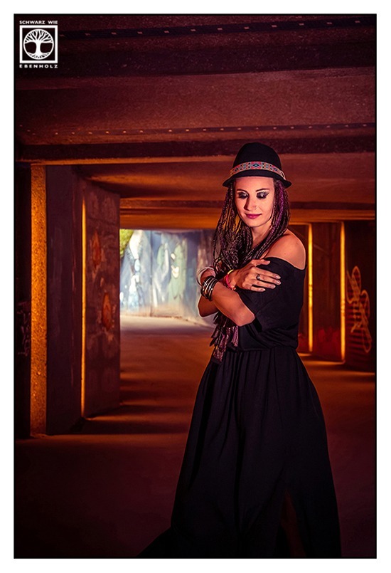 Boho Fotoshooting / Alternative Fotoshooting: Eine junge Frau steht seitlich in einer Unterführung. Sie umarmt sich selbst und blickt lächelnd zu Boden. Auf ihrem Kopf ist ein schwarzer Hut mit buntem Band im Ethno-Look. Sie hat lila und dunkelbraune Braids, die ihr bis zur Hüfte reichen und trägt ein langes, schwarzes Kleid mit kurzen Ärmeln, das ihr ein wenig von der Schulter gerutscht ist. An ihren Armgelenken sind Festivalbänder und silberne Reifen. Die Unterführung ist voller schöner Graffiti, die jeweils mit einem senkrechten Streifen aus Licht in Szene gesetzt werden.