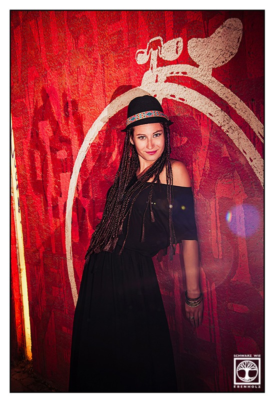 Boho Fotoshooting / Alternative Fotoshooting: Eine junge Frau steht vor einem orange-rotem Graffiti und wird von dem großen, weißen Rad eines gemalten Einrads eingerahmt. Sie lehnt mit ihrer gesamten Schulterpartie an der Wand, ihr linker Arm berührt diese komplett. Auf ihrem Kopf ist ein schwarzer Hut mit buntem Band im Ethno-Look. Sie hat lila und dunkelbraune Braids, die ihr bis zur Hüfte reichen und trägt ein langes, schwarzes Kleid mit kurzen Ärmeln und U-Boot-Ausschnitt. An ihrem Armgelenk sind silberne Reifen. Sie lächelt und blickt direkt in die Kamera.