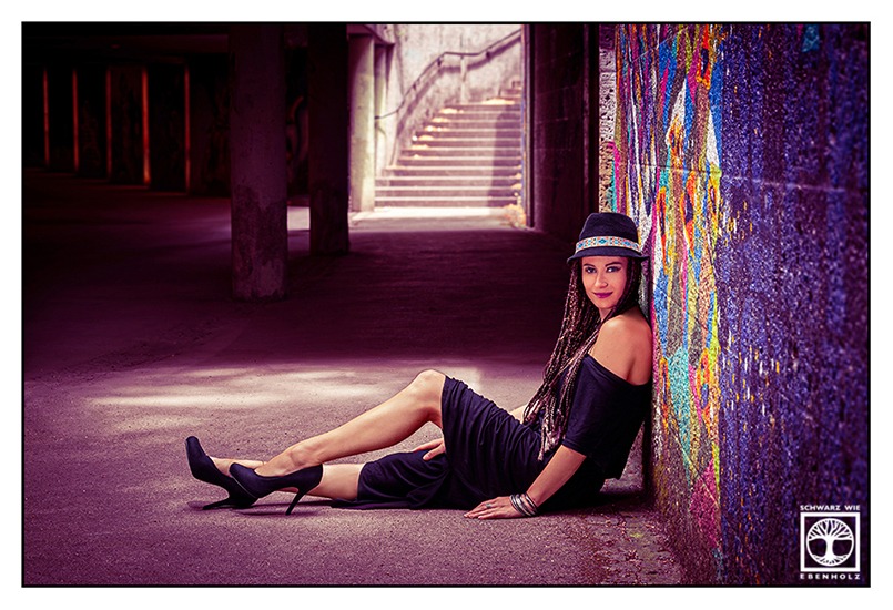 Boho Fotoshooting / Alternative Fotoshooting: Eine junge Frau sitzt in einer Unterführung vor einem farbenfrohen Graffiti. Ihre Beine sind zur linken Seite des Bildes ausgestreckt, ihr linkes, vorderes Bein steht angewinkelt auf dem Boden. Auf ihrem Kopf ist ein schwarzer Hut mit buntem Band im Ethno-Look. Sie hat lila und dunkelbraune Braids, die ihr bis zur Hüfte reichen und trägt ein langes, schwarzes Kleid mit kurzen Ärmeln und U-Boot-Ausschnitt. An ihrem Armgelenk sind silberne Reifen. Sie blickt uns direkt an. Im Hintergrund sieht man eine lichtüberflutete Treppe.