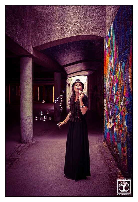 Boho Fotoshooting / Alternative Fotoshooting: Eine junge Frau steht frontal in einer Unterführung neben einem farbenfrohen Graffiti und direkt vor einer lichtüberfluteten Treppe. Sie macht gerade Seifenblasen. Ihr Augen sind geschlossen. Der Behälter mit der Seifenflüssigkeit ist in der rechten Hand und der Stab mit der runden Öffnung in der linken Hand. Auf ihrem Kopf ist ein schwarzer Hut mit buntem Band im Ethno-Look. Sie hat lila und dunkelbraune Braids, die ihr bis zur Hüfte reichen und trägt ein langes, schwarzes Kleid mit kurzen Ärmeln und U-Boot-Ausschnitt. An ihren Armgelenken sind Festivalbändchen und silberne Reifen.