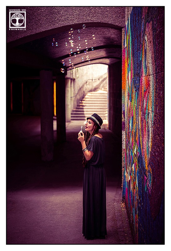 Boho Fotoshooting / Alternative Fotoshooting: Eine junge Frau steht seitlich in einer Unterführung neben einem farbenfrohen Graffiti und direkt vor einer lichtüberfluteten Treppe. Das Foto wurde jedoch leicht erhöht aufgenommen, so dass sie nur den unteren Teil der Treppe verdeckt. Sie macht gerade Seifenblasen. Ihr Augen sind geschlossen. Der Stab mit der runden Öffnung ist in der linken Hand. Auf ihrem Kopf ist ein schwarzer Hut mit buntem Band im Ethno-Look. Sie hat lila und dunkelbraune Braids, die ihr bis zur Hüfte reichen und trägt ein langes, schwarzes Kleid mit kurzen Ärmeln und U-Boot-Ausschnitt. An ihrem Armgelenk sind silberne Reifen.
