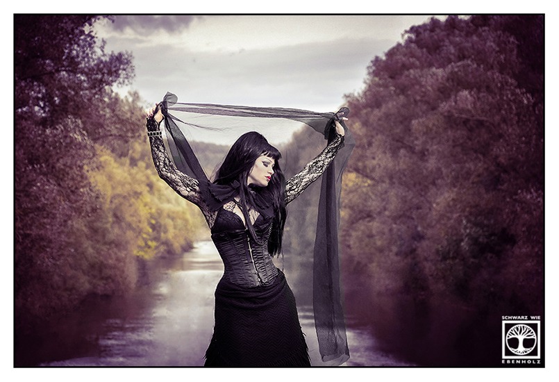 Gothic Shooting Outdoor: Ganz in schwarz gekleidet steht eine Gothic Lady vor einem von Bäumen gesäumten Fluss. Sie hält einen transparenten schwarzen Stoff hinter ihrem Kopf mit beiden Händen. Ihr Gesicht ist zur von uns aus rechten Seite gewandt. Sie hat lange schwarze Haare und trägt ein Korsett, Armstulpen aus Spitze und einen langen schwarzen Rock.