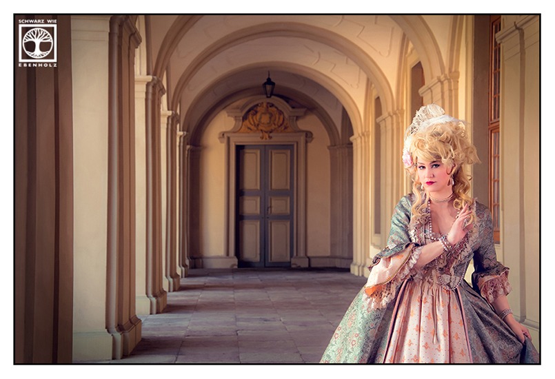 Barock Fotoshooting: Eine edle Frau mit blonden Locken, weißer Feder und rosa Blüten im Haar schreitet anmutig durch den Flur eines Schlosses. Sie trägt ein pompöses barockes Kleid in bläulichem Grün und hellem Orange sowie Perlenschmuck an Hals und Armen. Am Ende des Ganges ist eine blau umrandete Tür. An der von uns aus linken Seite sind Säulen und rechts. kann man Fenster erkennen.