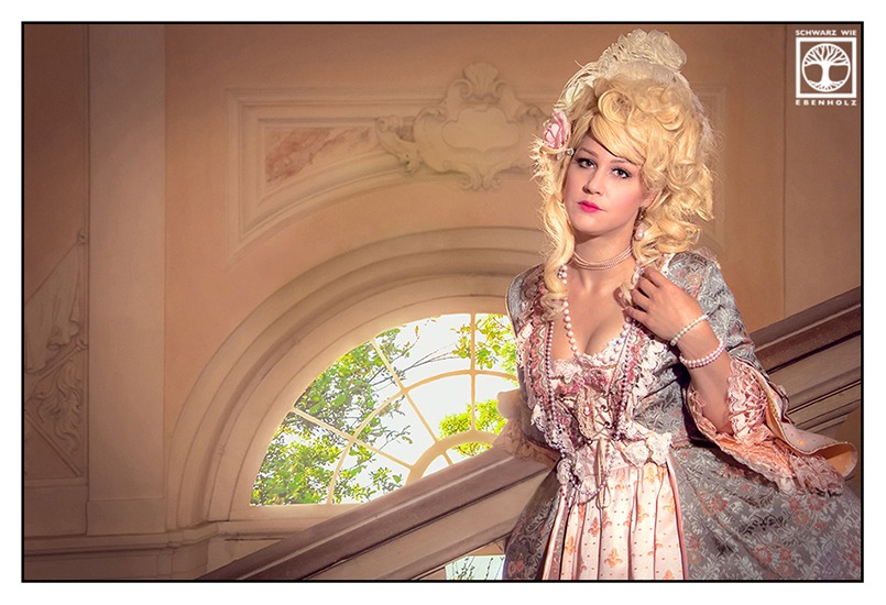 Fantasy Fotoshooting Marie Antoinette: Eine edle Dame steht im Schloss auf einer Treppe. Im Hintergrund ist ein halbrundes Fenster mit Bäumen zu sehen. Sie trägt eine hellblonde Lockenperücke, deren Haar locker hochgesteckt ist. Federn und eine rosa Stoffblume zieren das Haar. Um Hals und Armgelenke und an den Ohren baumelt Perlenschmuck. Das Kleid an sich ist apricot und hat ein eingearbeitetes Mieder. Darüber ist ein mintgrüner Überwurf.