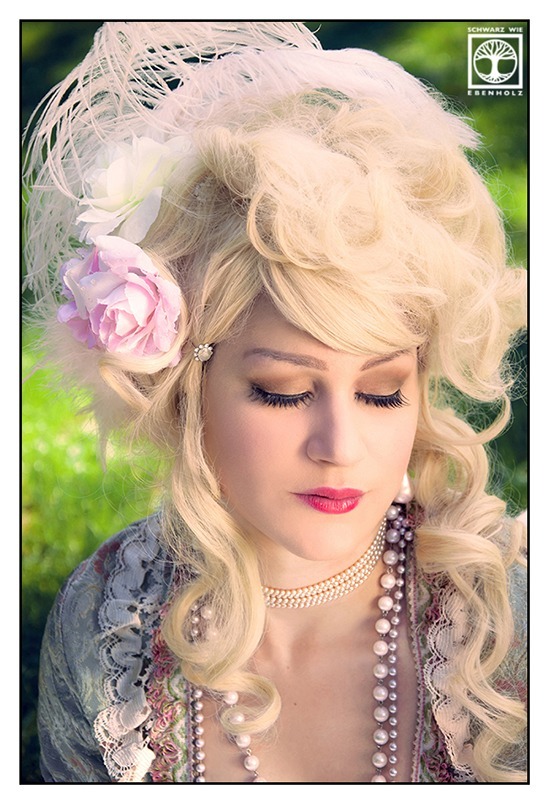 Fantasy Fotoshooting Marie Antoinette: Auf diesem Portrait ist eine edle Dame mit niedergeschlagenen Augen zu sehen. Ihr Kopf ist zur rechten Bildseite geneigt. Sie trägt eine hellblonde Lockenperücke, deren Haar locker hochgesteckt ist. Zwei Stoffblumen in rosa und weiß sowie zwei weiße Federn zieren das Haar. Mehrere Perlenketten hängen an ihrem Hals. Vom Kleid ist viel Spitze in weiß, rosa und mintgrün zu sehen.