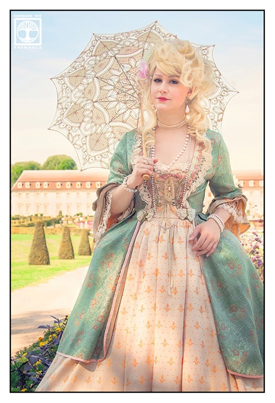 Fantasy Fotoshooting Marie Antoinette: Eine edle Dame in barockem Kleid und einem weißen Sonnenschirm aus Spitze steht anmutig vor einem Schloss mit Schlosspark, das sich klein im Hintergrund abzeichnet. Sie trägt eine hellblonde Lockenperücke, deren Haar locker hochgesteckt ist. Um Hals und Armgelenke und an den Ohren baumelt Perlenschmuck. Das Kleid an sich ist apricot und hat ein eingearbeitetes Mieder. Darüber ist ein mintgrüner Überwurf.