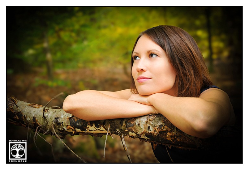 Natürliches Fotoshooting: Das Porträt seit eine brünette Frau mit schulterlangen Haaren und braunen Augen. Sie lehnt sich auf einen Ast und hat ihren Kopf auf den verschränkten Armen liegen. Sie blickt nach oben links und lächelt leicht. Im Hintergrund ist unscharf Wald zu erkennen.