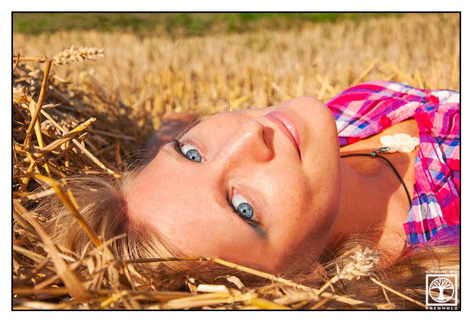 Fotoshooting Natur: Eine schlanke Frau mit langen blonden Haaren und kurzärmliger pinker Bluse mit Karomuster liegt in einem Stoppelfeld und bettet ihren Kopf auf Stroh. Sie lächelt und blickt uns an. Wir sehen sie auf Augenhöhe von oben.