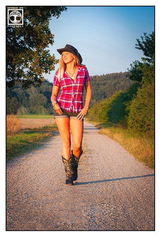 Fantasy Fotoshooting Cowgirl: Eine schlanke Frau mit langen blonden Haaren läuft auf einem Feldweg und schaut zur linken Seite des Bildes in Richtung Abendsonne. Sie trägt einen grauen Cowboyhut, eine pinke, kurzärmlige Bluse mit Karomuster, eine kurze blaugraue Jeans und Cowboystiefel. Ihre Daumen stecken in ihren Hosentaschen vorne.