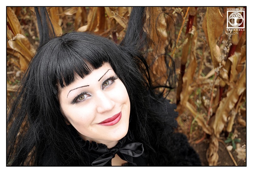 Beweisbild, dass ein Gothic lachen kann: Eine Gothic Lady im Dark Romantic Look (Düsterromantiker) sitzt in einem vertrockneten Maisfeld und grinst.